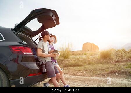 das asiatische Paar lehnte sich an der Rückseite des Autos und schaute auf die Aussicht, während es Kaffee an einem verlassenen historischen Ort trank Stockfoto
