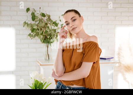 Eine junge, schöne Frau mit europäischem Aussehen in legerer Kleidung steht in einem Raum vor dem Hintergrund einer weißen Backsteinmauer, hinter ihr sind Blumen in einer Vase, die Sonnenstrahlen an den Wänden Stockfoto