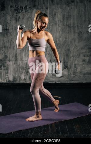 Portrait einer muskulösen jungen athletischen Frau mit perfektem schönen Körper, die Sportkleidung trägt und dabei Gewichte hebt und nach vorne lunging. Stockfoto