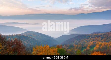 Herbstliche ländliche Landschaft. Wunderschöne Naturlandschaft mit nebligen Tal und glühenden Himmel bei Sonnenaufgang. Bäume in bunten Laub und Felder auf Hügeln am Morgen Stockfoto