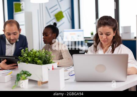 Fokussierte Managerin, die auf dem Laptop tippt, im Internet surft, während sie am Schreibtisch sitzt, konzentrierte sich auf Multitasking. Verschiedene Kollegen im Hintergrund. Multiethnische Mitarbeiter planen ein neues Finanzprojekt. Stockfoto