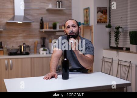 Nachdenklicher Mann, der ein Glas Wein trinkt, während er allein und deprimiert sitzt. Person mit Alkoholabhängigkeit mit einer Flasche alkoholisches Getränk, Schnaps, Alkohol, das Gefühl, heartbroken und verzweifelt Stockfoto