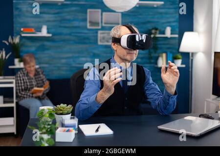 Glücklicher Rentner, der mit einem Headset die virtuelle Realität erlebt. Älterer Mann Rentner im Wohnzimmer sitzen am Schreibtisch mit futuristischen Brille für erweiterte Cyberspace Frau Fernsehen. Stockfoto