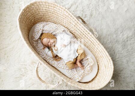 Ein neugeborenes Baby-Puppenspielzeug, gekleidet in einen senffarbenen Hut und eine weiße Strickjacke, die in einem cremefarbenen moses-Korb-Korb-Laderett liegt Stockfoto