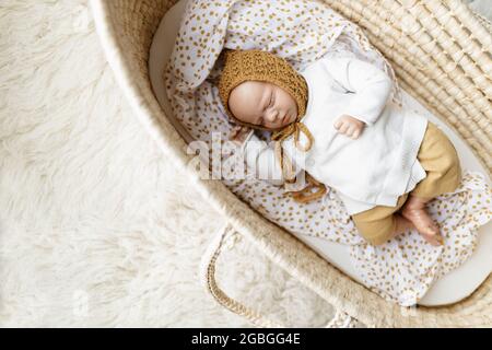 Ein neugeborenes Baby-Puppenspielzeug, gekleidet in einen senffarbenen Hut und eine weiße Strickjacke, die in einem cremefarbenen moses-Korb-Korb-Laderett liegt Stockfoto