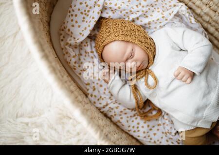 Ein neugeborenes Baby-Puppenspielzeug, gekleidet in einen senffarbenen Hut und eine weiße Strickjacke, die in einem cremefarbenen moses-Korb-Korb-Laderett liegt Stockfoto