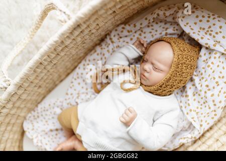 Ein neugeborenes Baby-Puppenspielzeug, gekleidet in einen senffarbenen Hut und eine weiße Strickjacke, die in einem cremefarbenen moses-Korb-Korb-Laderett liegt Stockfoto