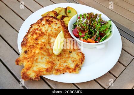 Wiener Schnitzel mit Zitronenscheibe, Salat und Bratkartoffel auf einem Holzgartentisch in einem Restaurant im Freien, ausgewählter Fokus, enge Schärfentiefe Stockfoto