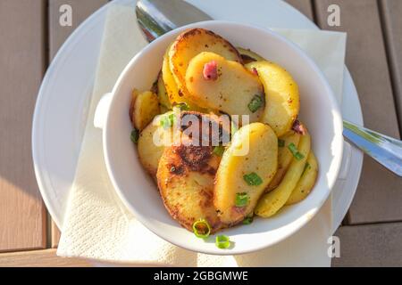 Gebratene Kartoffeln mit Speck und Lauch-Zwiebeln in einer Schüssel auf einem Holztisch bei einer Mahlzeit im Freien, Blick von oben, ausgewählter Fokus, enge Tiefe Stockfoto