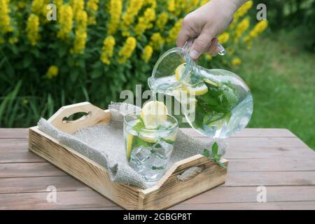 Eine Frauenhand gießt selbstgemachte Limonade in ein Glasglas, das auf einem Holztablett steht. Stockfoto