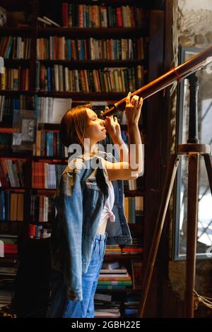 Die junge Hipster-Hündin schaut durch ein Teleskop, das im Vintage-Bibliotheksraum steht. Stockfoto