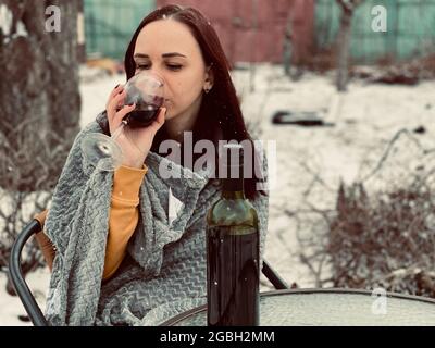 Junge Frau, die im Hof sitzt und bei verschneitem Wetter Rotwein trinkt. Frau in grauem Kariert gewickelt, die in der Wintersaison mit Alkohol auf der Straße sitzt Stockfoto