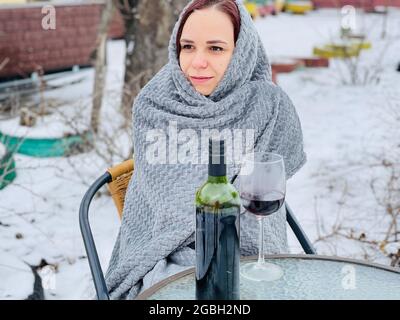 Junge Frau, die im Hof sitzt und bei verschneitem Wetter Rotwein trinkt. Frau in grauem Kariert gewickelt, die in der Wintersaison mit Alkohol auf der Straße sitzt Stockfoto