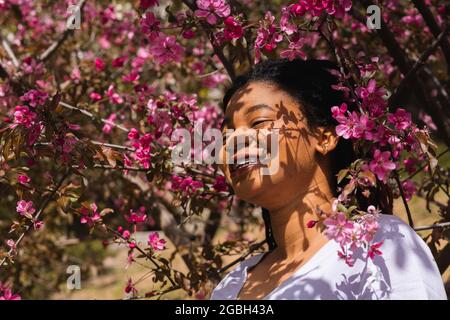 Porträt einer jungen glücklichen Afro-Frau in einem blühenden Rosenbaum mit Schatten und Sonnenlichtern auf ihrem Gesicht. Stockfoto