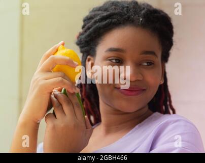 Junge dunkelhäutige Frau, die mit einer reifen Zitrone in den Händen posiert, lächelt sie Stockfoto