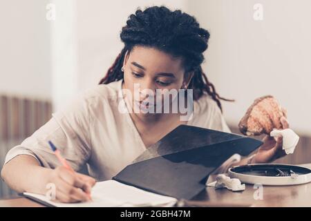 Junge Afro-Studentin schreibt in einem Tablet, während sie in einem Café zum Mittagessen sitzt und hält ein Croissant in der anderen Hand. Farblich abgestuft Stockfoto