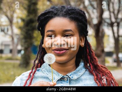 Porträt einer jungen, glücklichen Afro-Frau, die einen Dandelion in der Hand vor ihrem Gesicht hält Stockfoto