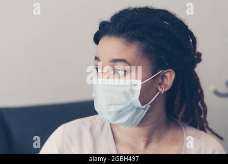 Eine afroamerikanische Frau sitzt vor dem Hintergrund einer hellen Wand. Auf dem Gesicht einer Person eine schützende medizinische Maske. Keine Emotionen Stockfoto