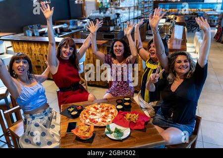 Eine Gruppe reifer Frauen, die in einem Pub sitzen, heben ihre Hände, während sie Pizza essen und Bier trinken Stockfoto