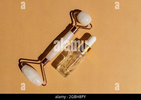 Massagerolle für das Gesicht aus Rosenquarz mit einer Flasche 24k Gold Serum auf beigem Hintergrund. Das Konzept der Hautpflege zu Hause Stockfoto