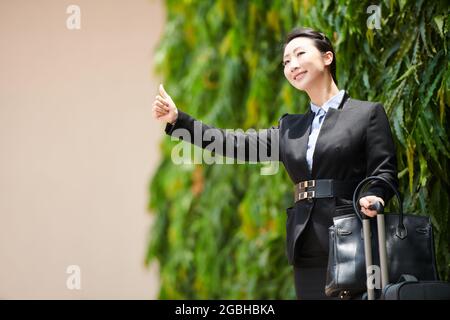 Lächelnde junge asiatische Geschäftsfrau in schwarzer formalwear, die mit Koffer steht und beim Taxi-Fang Daumen hoch zeigt Stockfoto