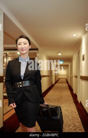Contetn Attraktive junge asiatische Frau in formalwear, die auf Geschäftsreise mit Gepäck durch den Hotelkorridor läuft Stockfoto