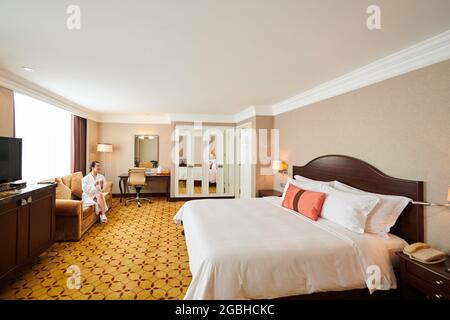 Frau im Bademantel sitzt auf dem Sofa und trinkt Kaffee in einem großen Hotelzimmer mit Doppelbett Stockfoto