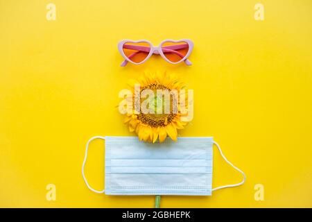 Lustiges Gesicht aus rosa herzförmiger Sonnenbrille, einer schützenden medizinischen Maske und einer Sonnenblume auf hellgelbem Hintergrund Stockfoto