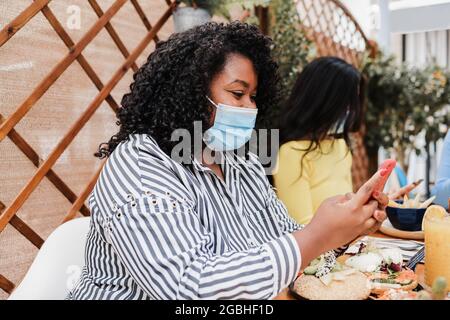 Fröhliche, multirassische Freunde, die im Brunch-Restaurant im Freien Handys benutzen und dabei Masken tragen - konzentrieren Sie sich auf afroamerikanische Mädchen Stockfoto