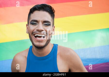 Junger Transgender-Mann mit Make-up lächelt vor der Kamera mit lgbt-Regenbogenfahne auf dem Hintergrund - Fokus auf Gesicht Stockfoto