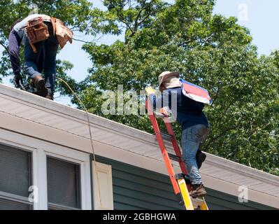 Auftragnehmer klettert Leiter mit Dachschindeln, während ein anderer Nagel die Schindeln auf ein Wohnhaus. Stockfoto