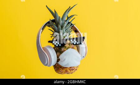 Kreative lustige Ananas Gesicht trägt Brille Kopfhörer und schützende medizinische Maske. Ruhende schwebender Ananas Gesicht auf Farbe gelb Sommer Stockfoto