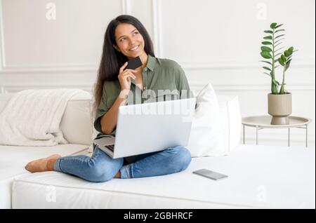 Bequemlichkeit zum Einkaufen, ohne das Haus zu verlassen. Die träumende inderin hält die Kreditkarte, die zu Hause mit einem Laptop auf dem Schoß auf der Couch sitzt, Einkäufe tätigt, Essen bestellt, nachdenklich zur Seite schaut Stockfoto