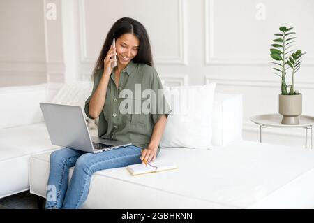 Multitasking indische freiberufliche Frau spricht auf dem Smartphone und nimmt Notizen mit Stift, mit Laptop für Remote-Arbeit von zu Hause, notieren einige Informationen sitzen auf dem Sofa, biracial Mädchen arbeitet aus der Ferne Stockfoto