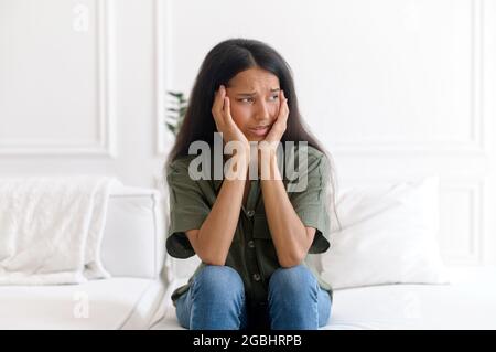 Eine verängstigte inderin fühlt sich in Panik versetzt, sitzt zu Hause auf dem Sofa, hält den Kopf fest und blickt mit Angst-Gesichtsausdruck davon. Verärgert Mischlinge Dame hat Problem und Depression, hat schlechte Gesundheit, Angst Stockfoto