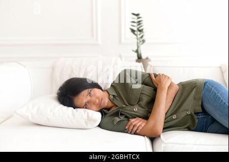 Deprimierte junge inderin mit dunklen Haaren, die mit gekreuzten Armen auf der Couch liegt, überwältigt, gestresst wegen unerfüllter Liebe oder aufbricht, nachdem sie mit den Eltern gekämpft hat Stockfoto