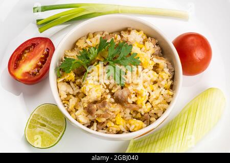 Draufsicht auf köstlichen hausgemachten thai-Stil Schweinefleisch und Ei gebratenen Reis in Keramikschale auf weißem Keramikplatte mit ganz und halb Tomaten, Gurken in Scheiben geschnitten, Stockfoto