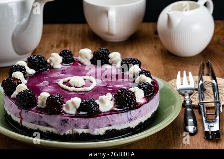 Seitenansicht des hausgemachten Brombeer-Käsekuchen, garniert mit konservierter Backberry und weißer Creme in grüner Keramikplatte auf Holztisch mit einer Tasse CO Stockfoto