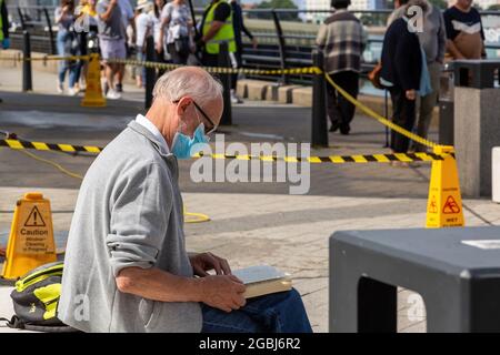 08-04-2021 Portsmouth, Hampshire, Großbritannien EIN Mann, der draußen auf einer Bank sitzt und eine Gesichtsmaske trägt, während er ein Buch liest Stockfoto