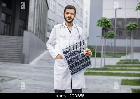 Hübscher arabischer Arzt mit Stethoskop, mit weißem Mantel, lächelnd auf die Kamera schauend, Tomographie haltend, draußen in der Nähe der Klinik stehend. Medizinisches und radiologisches Konzept Stockfoto