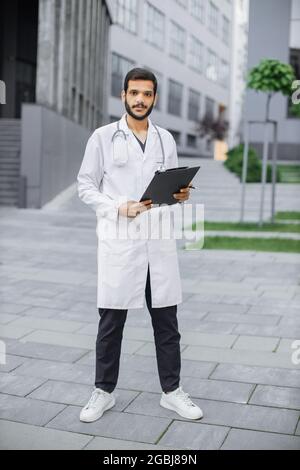 In voller Länge Porträt eines jungen, hübschen bärtigen arabischen Arztes im weißen Mantel, der mit einem Klemmbrett-Ordner in den Händen im Freien vor einem modernen Krankenhaus oder einer Klinik steht Stockfoto