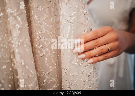 Nahaufnahme der Hand der Braut mit einem Ring darauf, der ihr wunderschönes Hochzeitskleid berührt Stockfoto