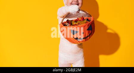 Kind im Mumienkostüm mit Pralinenkorb vor gelbem Hintergrund. Halloween Trick or Treat Stockfoto