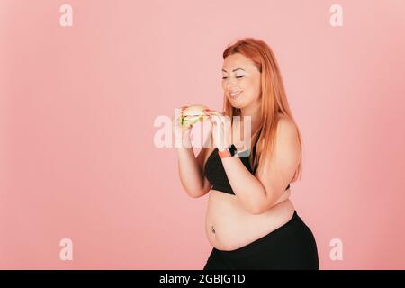 Fröhliche fröhliche plus size Frau trägt sportliche Outfit essen Fast Food Burger, während sie auf rosa Studio Hintergrund stehen. Stockfoto