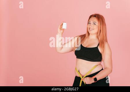 Schöne lächelnde übergroße Frau trägt sportliche Outfit Messung ihrer Taille und hält Glas von Pillen, während auf rosa Hintergrund stehen, kopieren Raum. Stockfoto