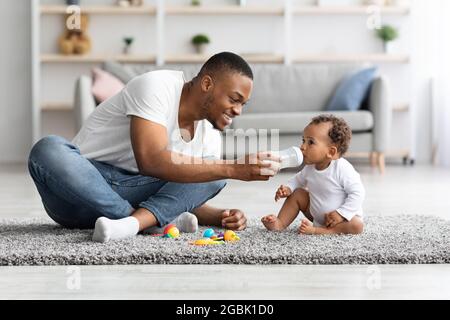 Fürsorglicher junger schwarzer Vater, der seinem kleinen Baby zu Hause Wasser gibt Stockfoto