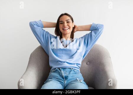 Junge Frau, die im Sessel sitzt, die Hände hinter dem Kopf hält und sich auf weißem Hintergrund entspannt Stockfoto