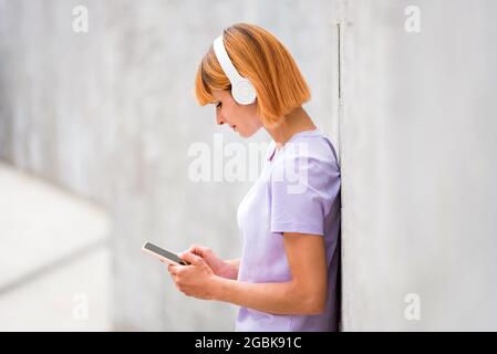 Attraktive Rothaarige, die Musik auf Stereo-Kopfhörern hört und auf ihrem Mobiltelefon nach Soundtracks surft, während sie sich gegen eine weiße Wa lehnt Stockfoto