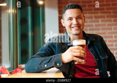 Glücklicher, attraktiver hispanischer Mann, der in einer Kneipe oder Bar ein kaltes Bier genießt und sein Glas hochhebt und der Kamera ein freundliches Lächeln schenkt Stockfoto