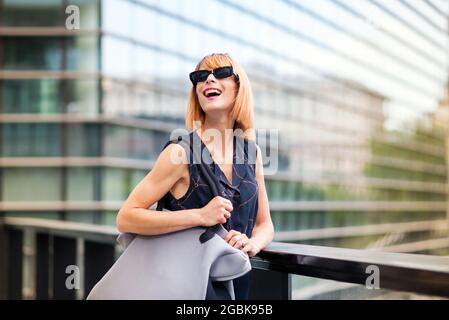 Lebhaft trendige Frau in moderner Sonnenbrille, die in einer Stadt gegen das Geländer eines Gehwegs steht und fröhlich lacht, während sie mit Glas in den Himmel blickt Stockfoto